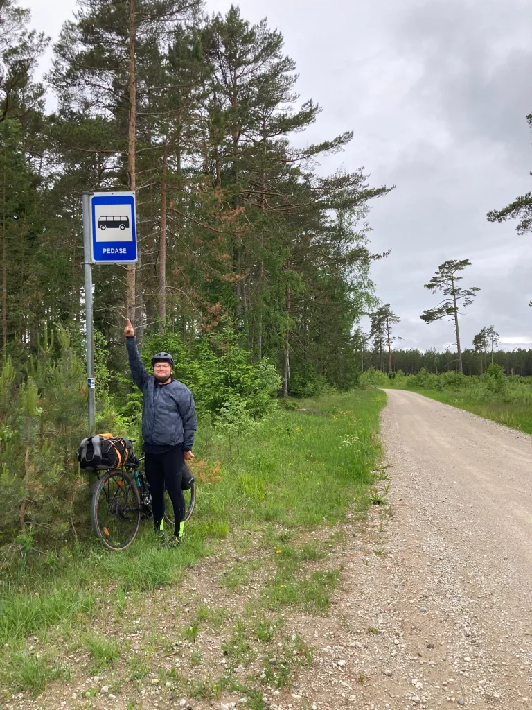 A cyclist with a loaded bike points to a Pedase bus stop sign on a rural gravel road