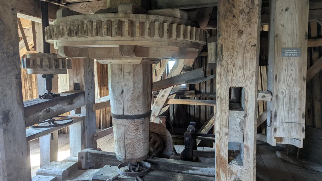 Large wooden gears and a central vertical shaft inside a traditional mill with a flour tube chute
