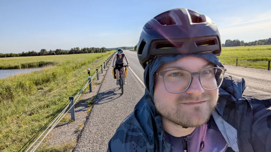 A man in a helmet takes a selfie while cycling on a path, with a woman following behind him on her bike