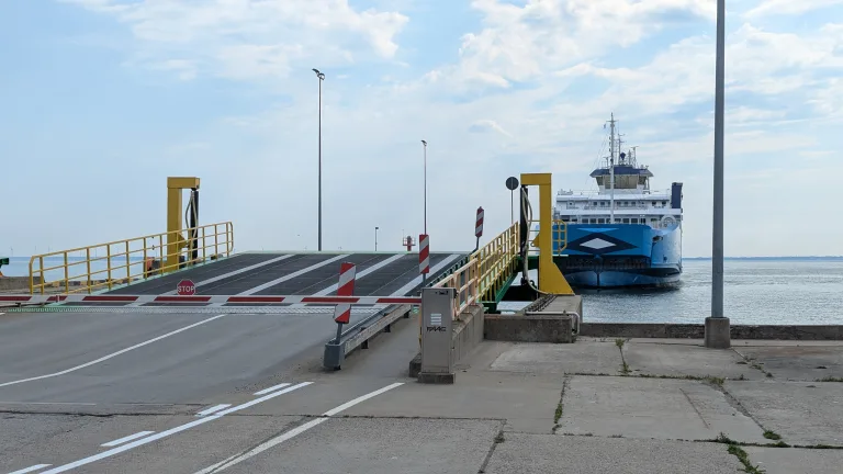 A blue and white ferry ship approaching a closed loading ramp at a harbor