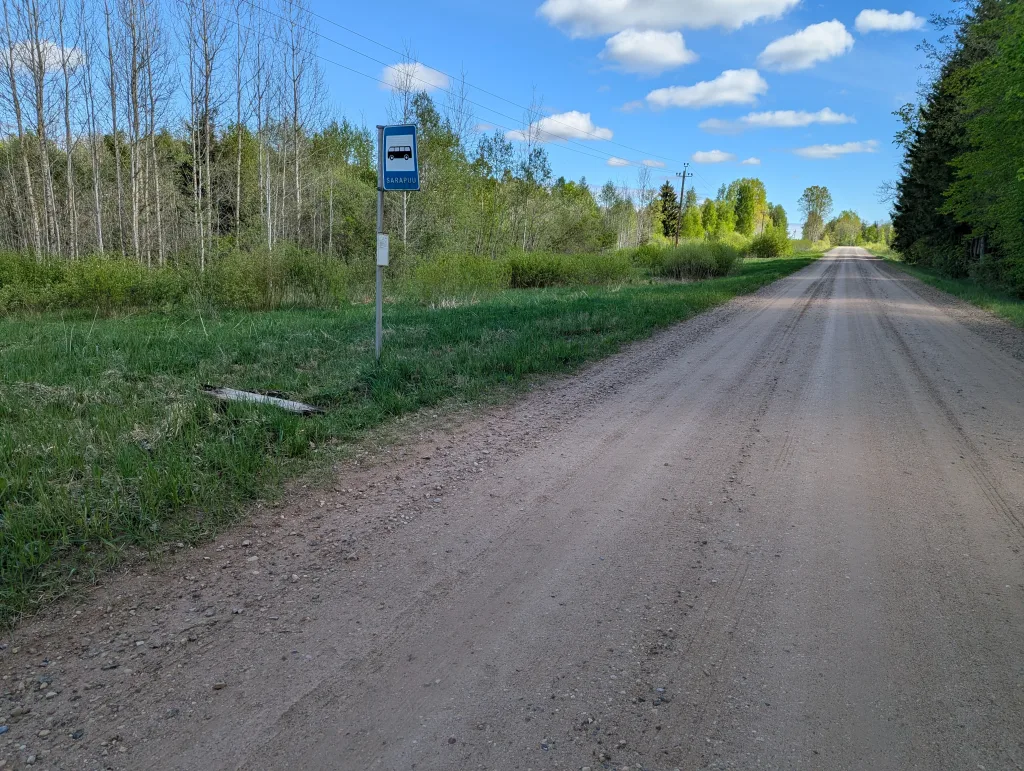 A wide, straight gravel road through rural countryside, with a blue bus stop sign labeled Sarapuu on the roadside