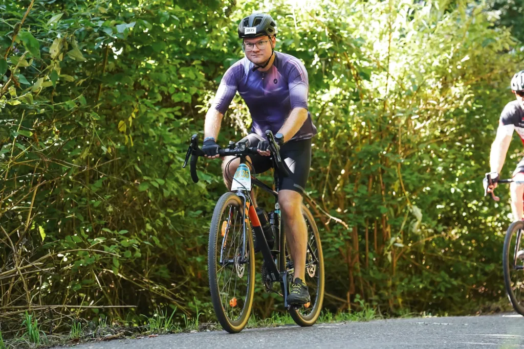 A cyclist in a purple jersey riding along a paved road surrounded by lush greenery, followed by another cyclist